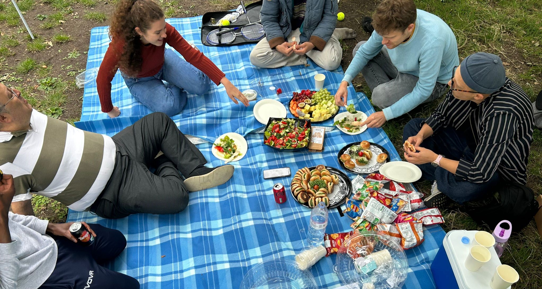 HIAS+JCORE staff, JUMP befrienders and young people sit on a blanket at the 2023 JUMP summer picnic. They are talking and eating food together.
