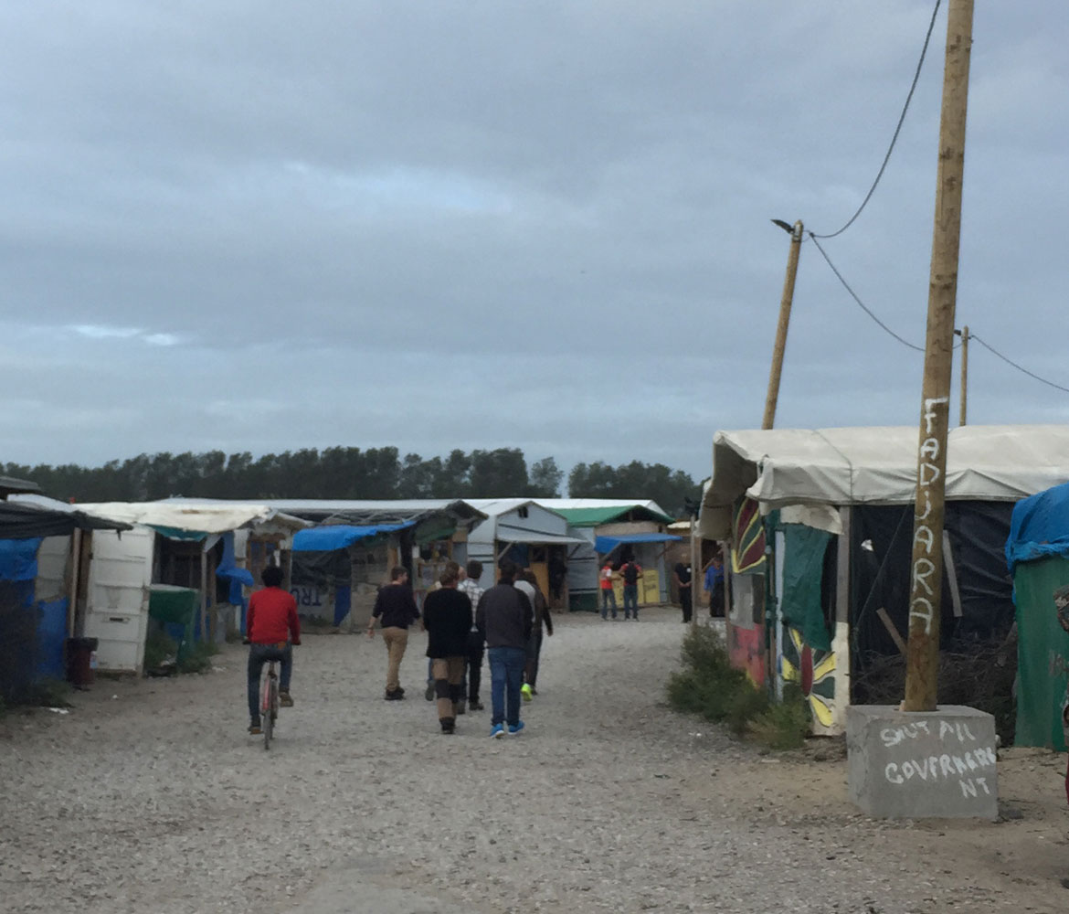 A refugee camp in Europe. A group of men are walking through the camp with their backs turned to the camera.